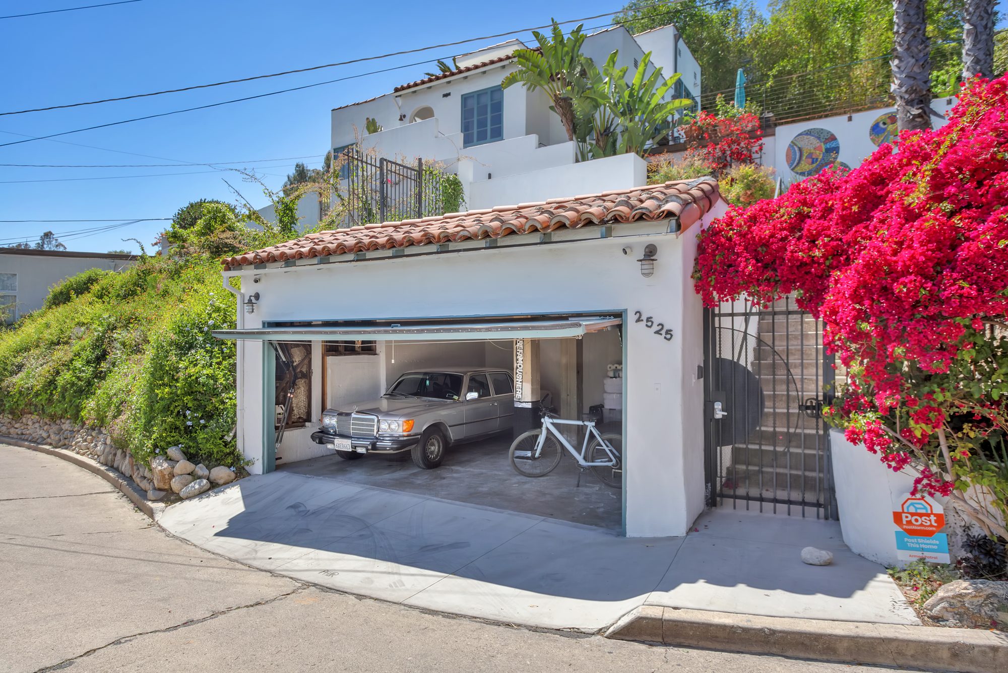 The two-car garage at street level with a clay tile roof, the number 2525, an iron gate to the entry stair, and a mass of magenta bougainvillea