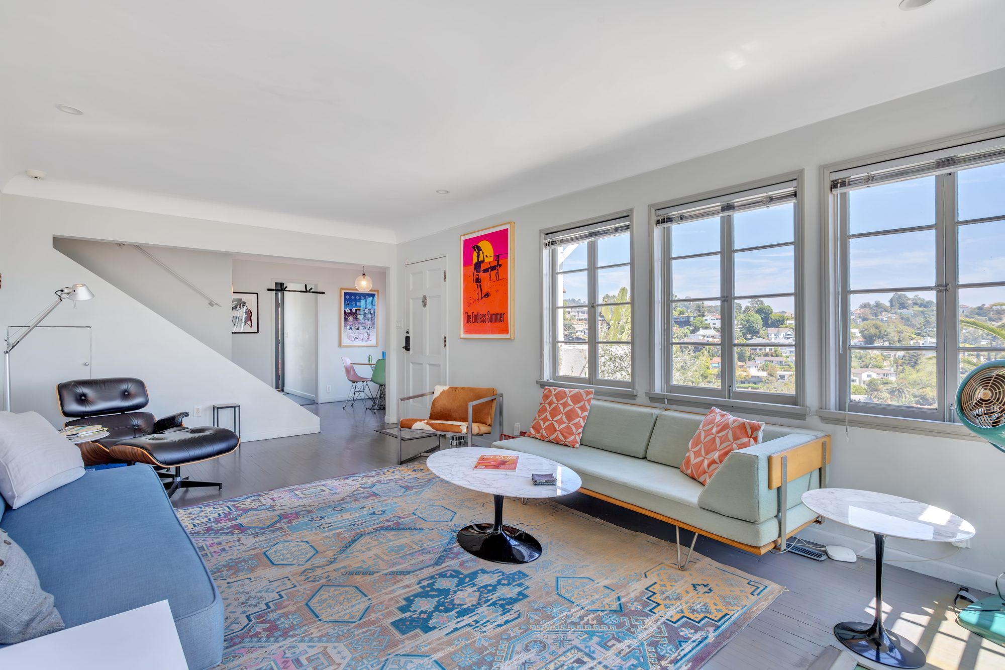 Living room with three wood casement windows looking over Beachwood Canyon rooftops, hardwood floors, and a stair rising to the bedroom level