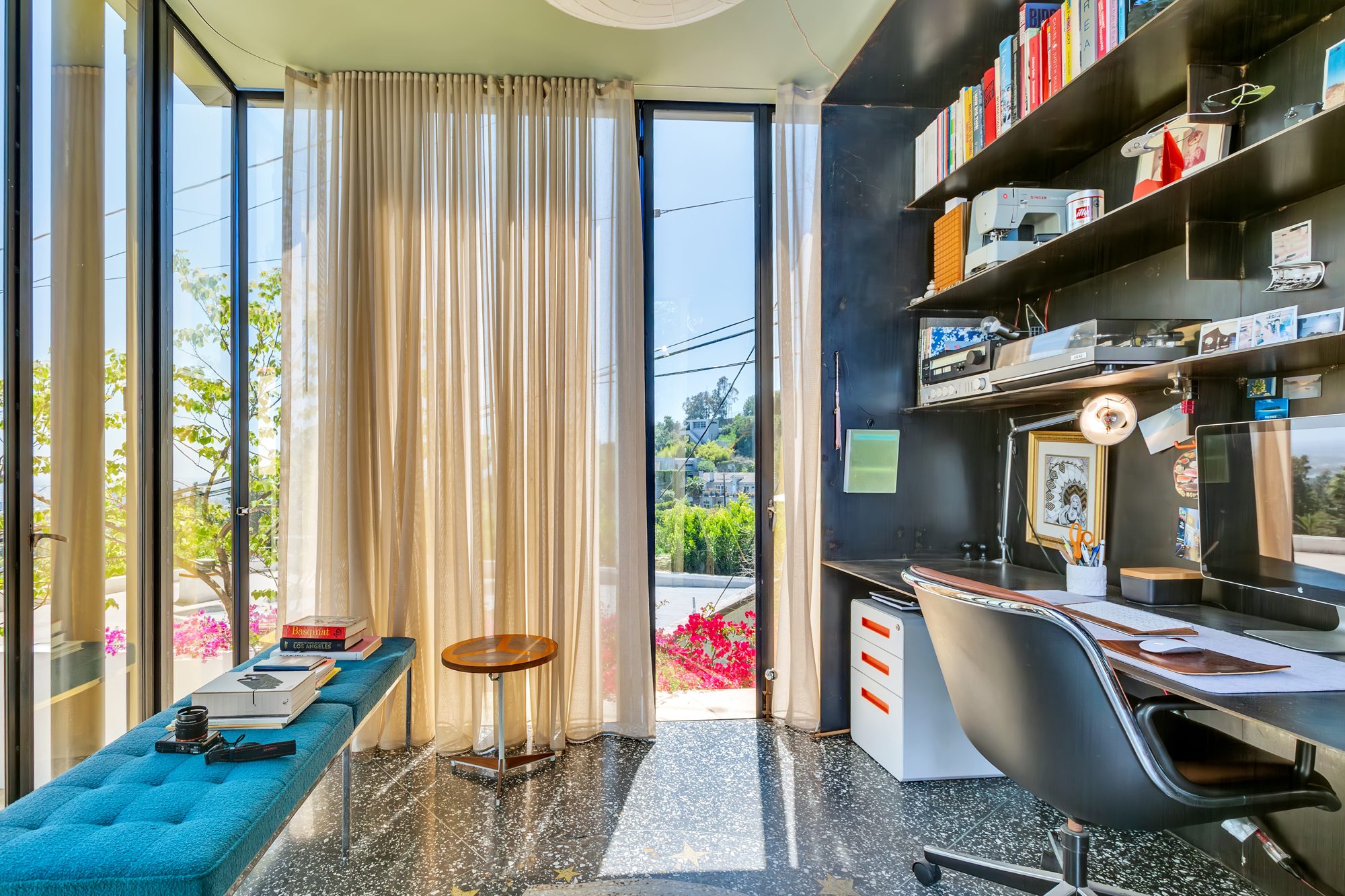 Inside the studio: a black steel desk and bookshelf wall, a terrazzo floor, sheer curtains, and glass walls open to the garden