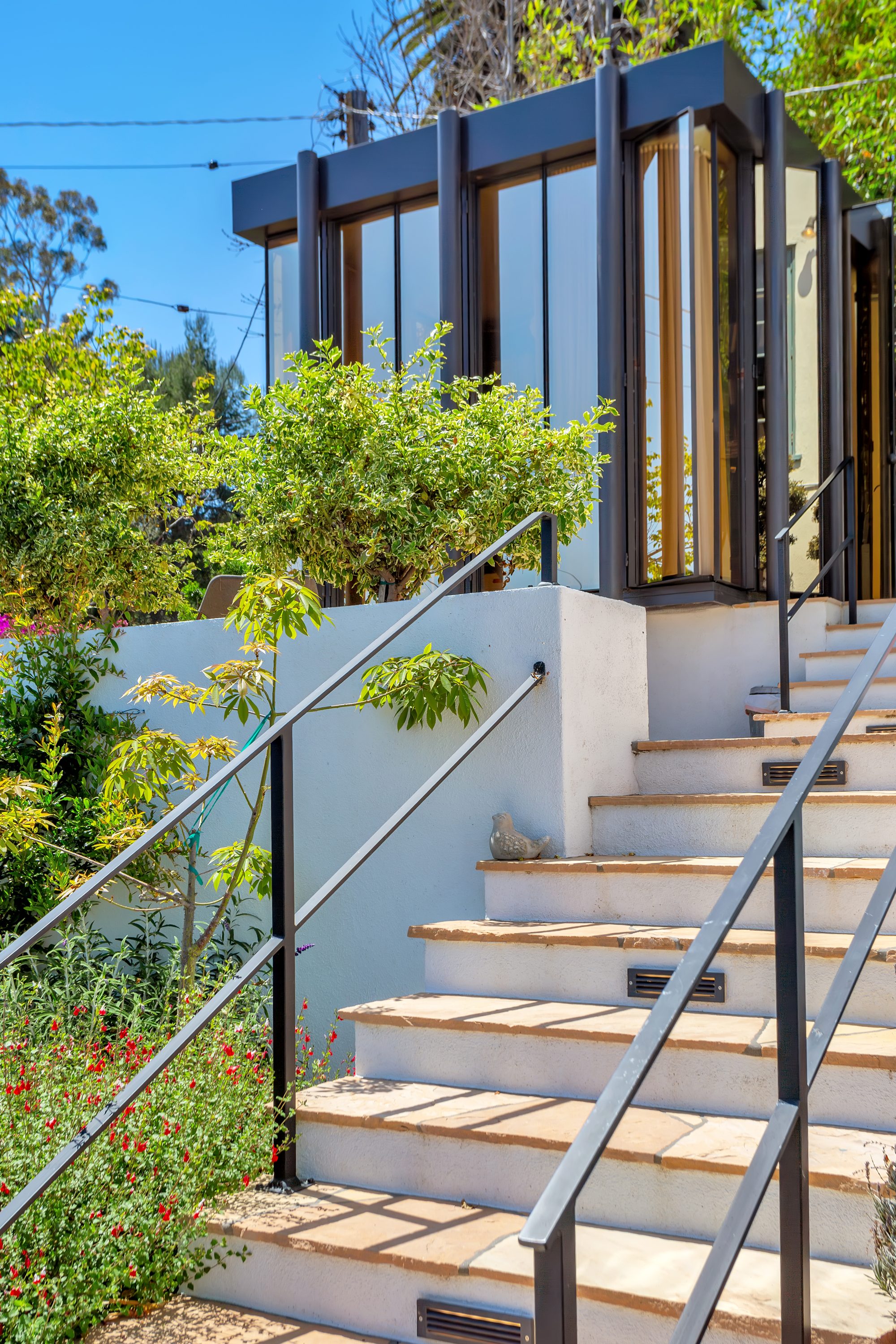 Stone steps with a black steel handrail rising to the glass studio, bordered by flowering plants