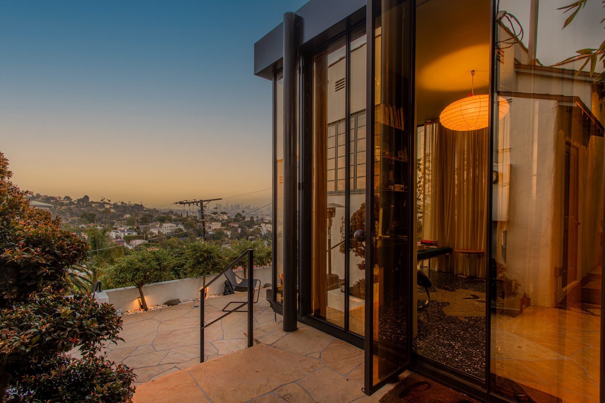 The steel-and-amber-glass studio lit from within at dusk on a flagstone terrace, with the downtown Los Angeles skyline in the distance